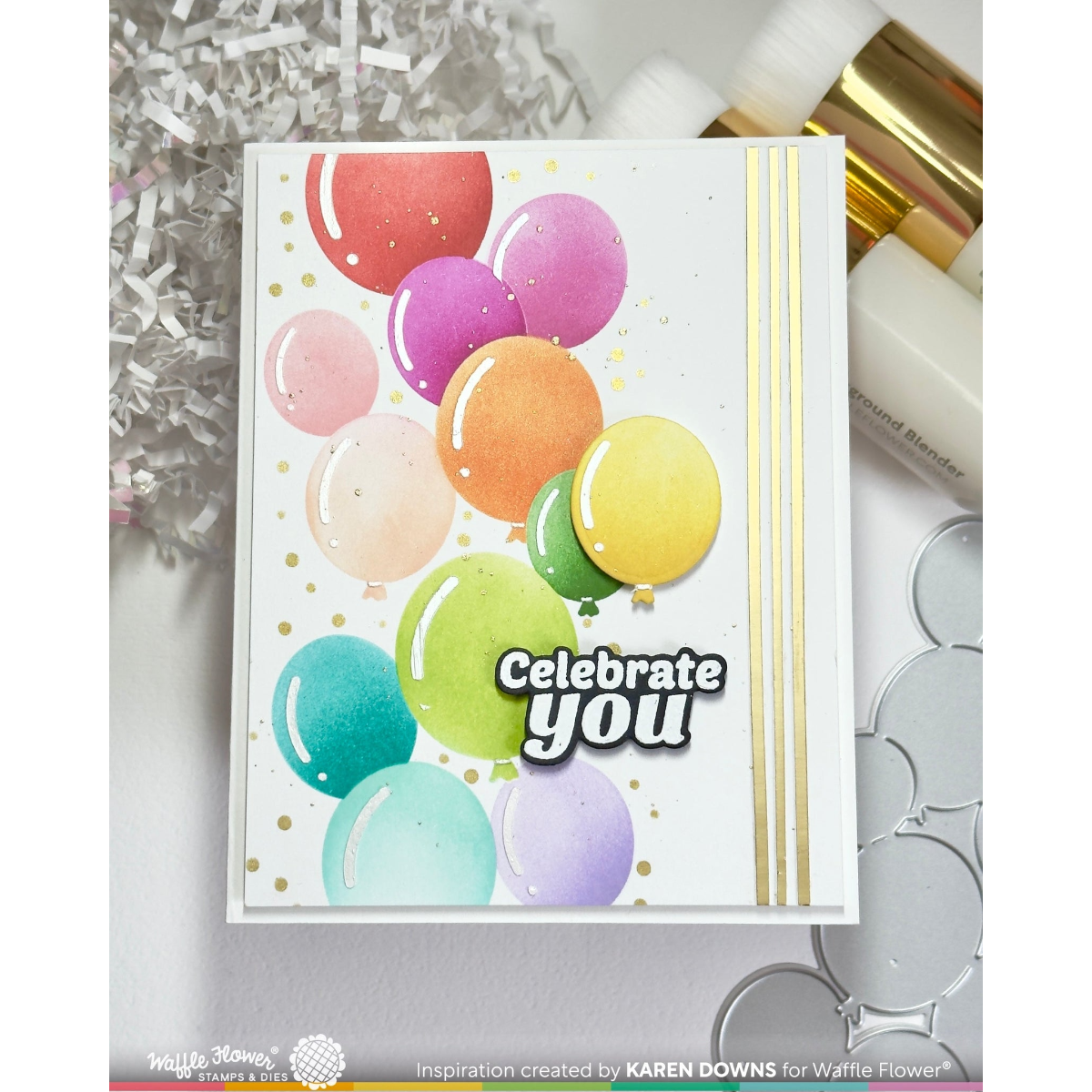 A handmade greeting card featuring colorful watercolor balloons, gold foil dots, and the text 'Celebrate you' in white with black outlines. The card is displayed on a light gray surface with craft supplies in the background.