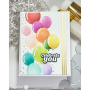 A handmade greeting card featuring colorful watercolor balloons, gold foil dots, and the text 'Celebrate you' in white with black outlines. The card is displayed on a light gray surface with craft supplies in the background.