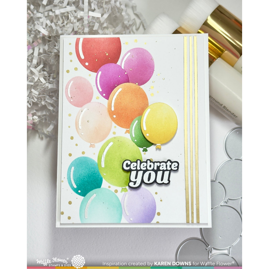 A handmade greeting card featuring colorful watercolor balloons, gold foil dots, and the text 'Celebrate you' in white with black outlines. The card is displayed on a light gray surface with craft supplies in the background.