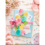 A handmade square birthday card with a light blue base, decorated with colorful balloon shapes in pink, yellow, green, and blue, a pink ribbon bow, and a white die-cut outline. The card is styled on a distressed white wood surface with pink flowers, a pink jar, and a lace doily.