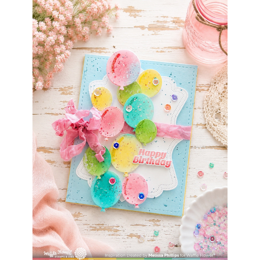 A handmade square birthday card with a light blue base, decorated with colorful balloon shapes in pink, yellow, green, and blue, a pink ribbon bow, and a white die-cut outline. The card is styled on a distressed white wood surface with pink flowers, a pink jar, and a lace doily.