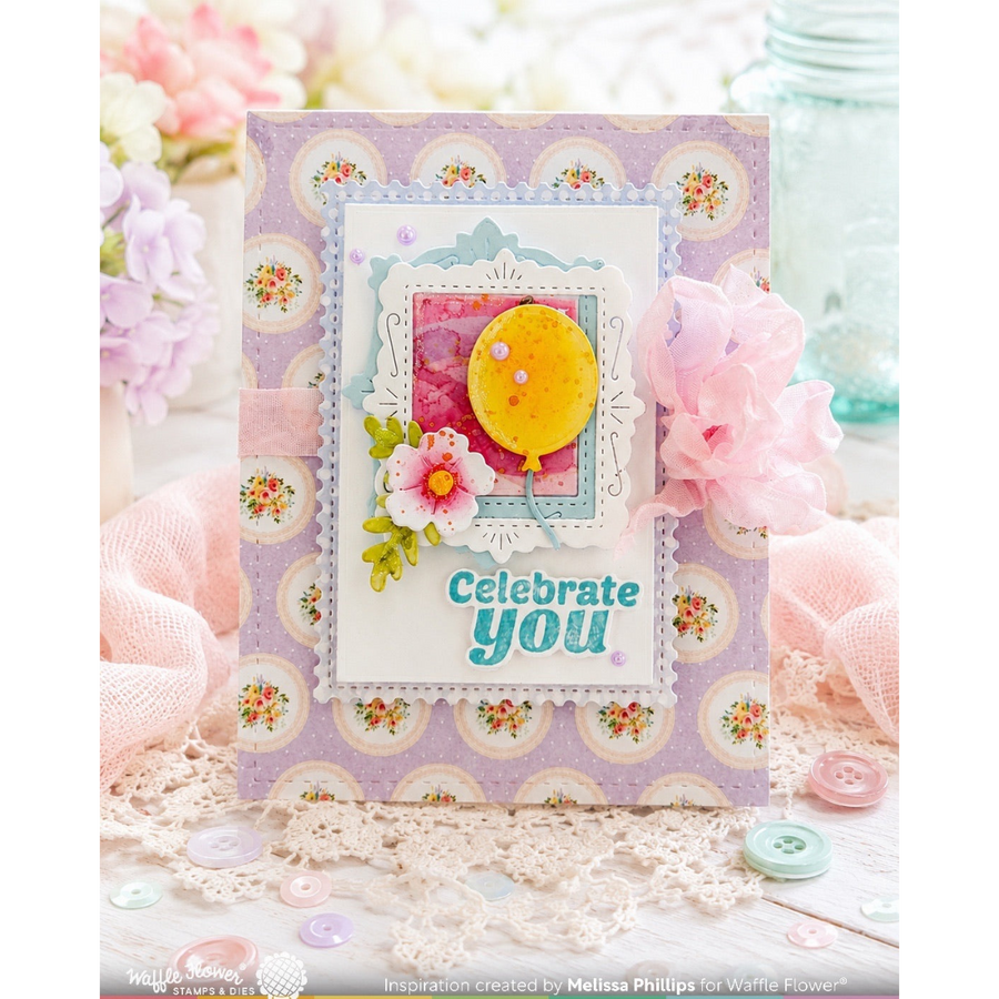 A handmade greeting card with a lavender floral border and a central layered design featuring a yellow balloon, pink flower, and the words 'Celebrate you' in blue. The card is styled on a lace surface with pink fabric, buttons, and blurred flowers in the background.