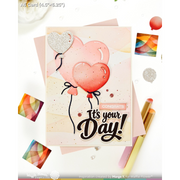 A handmade greeting card featuring two heart-shaped balloons, one pink and one silver glitter, with the text 'It's your Day!' and 'CONGRATS'. The card is styled on a white background with pink envelopes, colorful square cards, and gold pens.