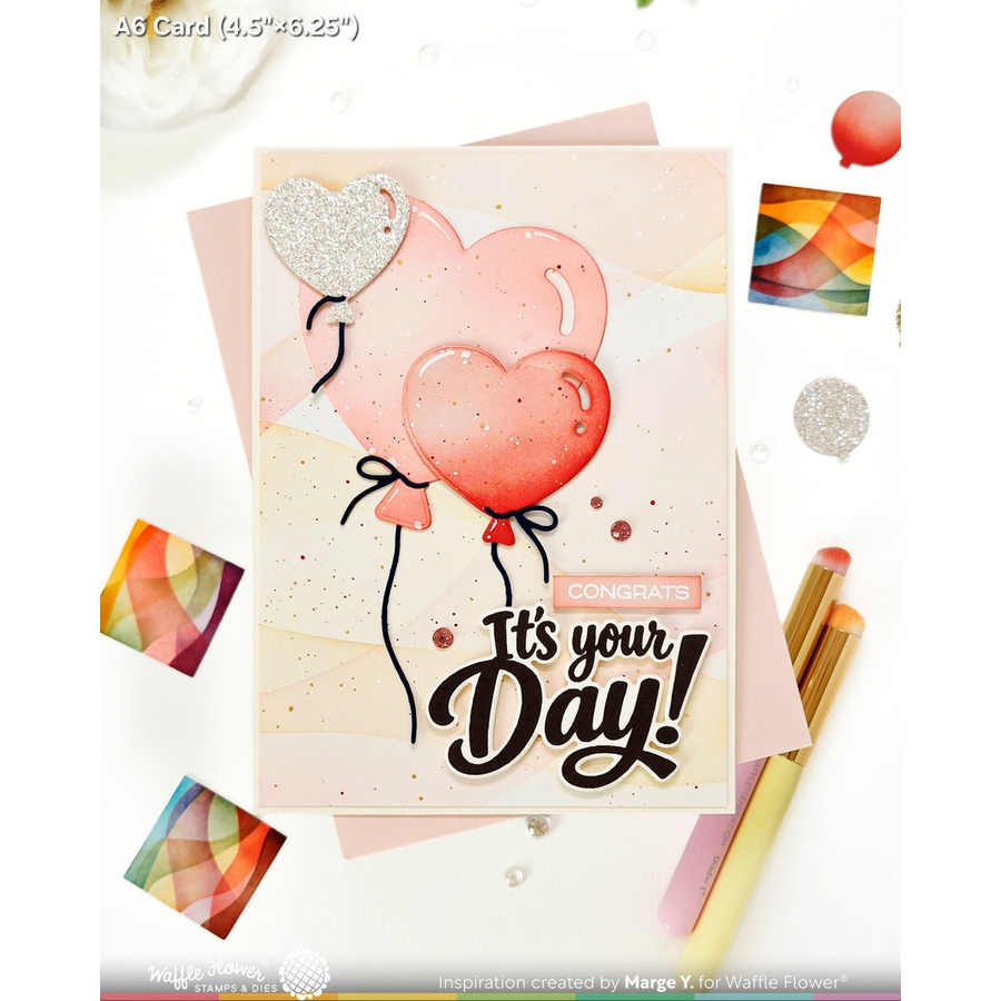 A handmade greeting card featuring two heart-shaped balloons, one pink and one silver glitter, with the text 'It's your Day!' and 'CONGRATS'. The card is styled on a white background with pink envelopes, colorful square cards, and gold pens.