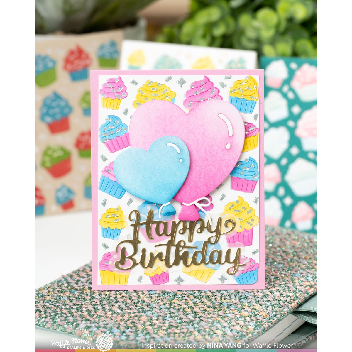 A handmade layered card featuring two heart-shaped balloons in pink and blue, surrounded by colorful cupcakes, with 'Happy Birthday' written in metallic gold script. The card is displayed on a speckled surface with blurred craft supplies in the background.