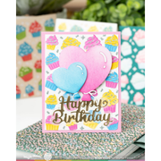 A handmade layered card featuring two heart-shaped balloons in pink and blue, surrounded by colorful cupcakes, with 'Happy Birthday' written in metallic gold script. The card is displayed on a speckled surface with blurred craft supplies in the background.