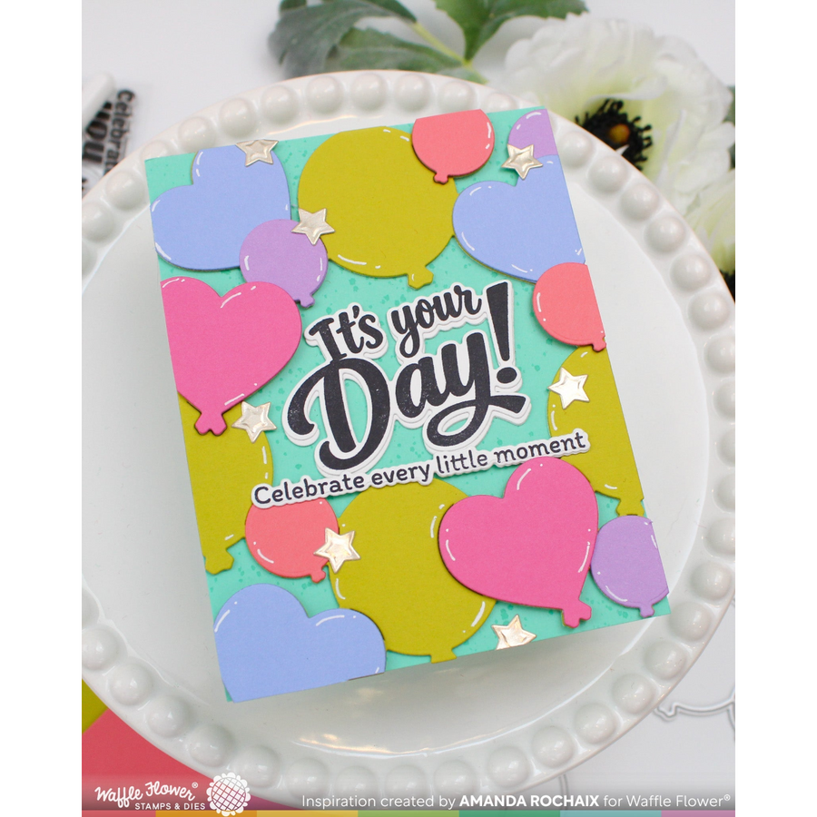A handmade greeting card featuring a colorful balloon and heart pattern with the text 'It's your Day! Celebrate every little moment', displayed on a white beaded plate.