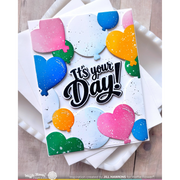 A handmade greeting card with layered heart-shaped balloon cutouts in pink, blue, green, and orange, featuring the text 'It's your Day!' in black script with white outlines. The card is displayed on top of white envelopes on a light wood surface.