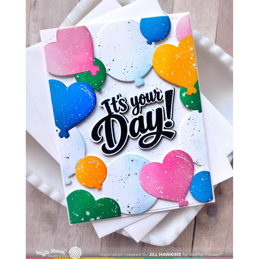 A handmade greeting card with layered heart-shaped balloon cutouts in pink, blue, green, and orange, featuring the text 'It's your Day!' in black script with white outlines. The card is displayed on top of white envelopes on a light wood surface.