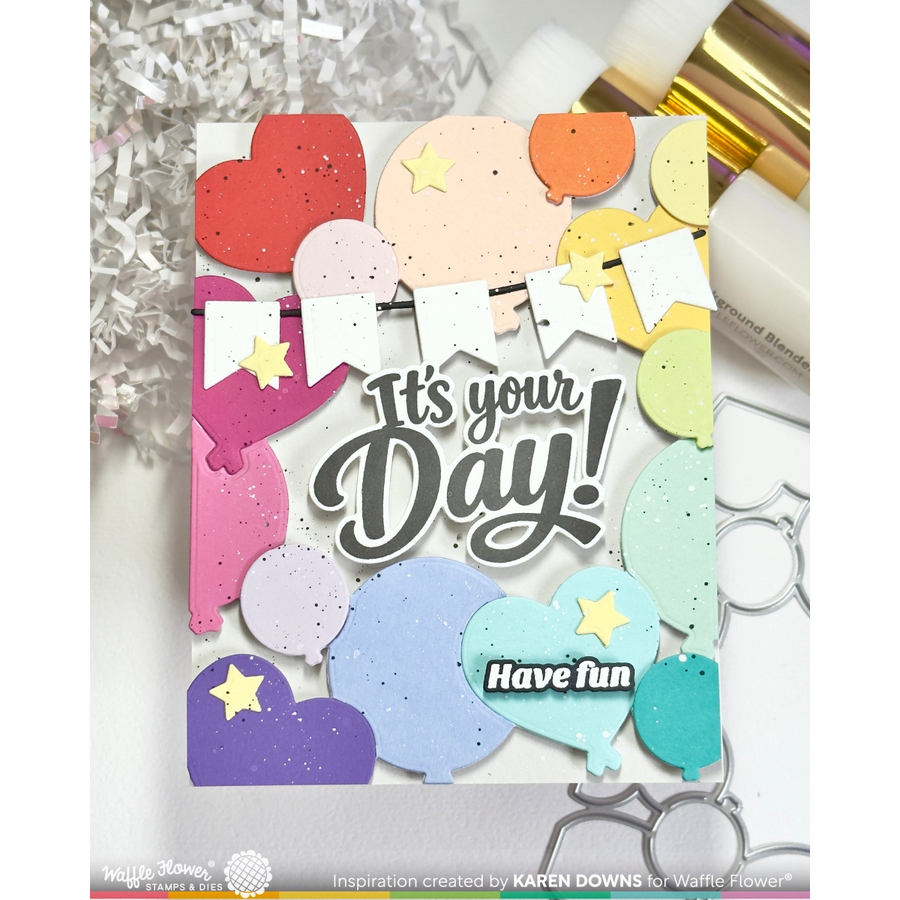 A handmade greeting card featuring colorful die-cut balloon shapes, bunting flags, and star accents with the text 'It's your Day! Have fun'. The card is displayed on a white surface with crafting tools and silver foil in the background.