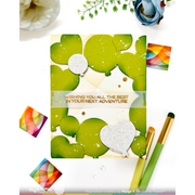 A handmade greeting card with green heart-shaped cutouts, a silver glitter heart, and the text 'WISHING YOU ALL THE BEST IN YOUR NEXT ADVENTURE' in gold foil. The card is styled in a flat-lay arrangement with craft brushes, small colorful paper swatches, and flowers on a white background.