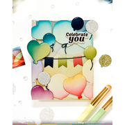 A square handmade greeting card or art print featuring layered heart-shaped balloons in pastel and bright colors, a banner, and the words 'Celebrate you.' The card is styled on a white background with scattered confetti, bubbles, and two paintbrushes in the foreground.