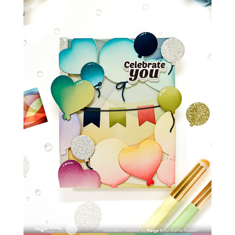 A square handmade greeting card or art print featuring layered heart-shaped balloons in pastel and bright colors, a banner, and the words 'Celebrate you.' The card is styled on a white background with scattered confetti, bubbles, and two paintbrushes in the foreground.