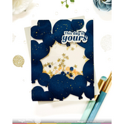 A handmade layered card featuring navy blue balloon shapes with gold glitter, a white central insert, and a 'This is yours' sticker. The card is styled on a white background with decorative pens, glitter, and flowers.