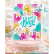 A handmade greeting card displayed on a pink pedestal, featuring colorful watercolor balloons, bunting flags, and the phrase 'It's your Day!' in teal script. The card is styled in a bakery-themed setting with cupcakes and a glass jar in the background.