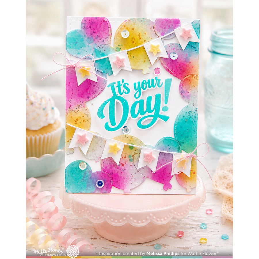 A handmade greeting card displayed on a pink pedestal, featuring colorful watercolor balloons, bunting flags, and the phrase 'It's your Day!' in teal script. The card is styled in a bakery-themed setting with cupcakes and a glass jar in the background.