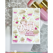 A handmade greeting card featuring colorful cupcake illustrations and the text 'It's your Day! Happy Birthday' in pink and white. The card is styled in a flat-lay arrangement with cosmetic brushes, silver tinsel, and a metal stamp die set on a white surface.