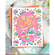 A handmade greeting card featuring colorful cupcakes and the text 'It's your Day! enjoy it' inside a pink oval, displayed on an orange cardstock sheet with crafting tools and yarn around it.