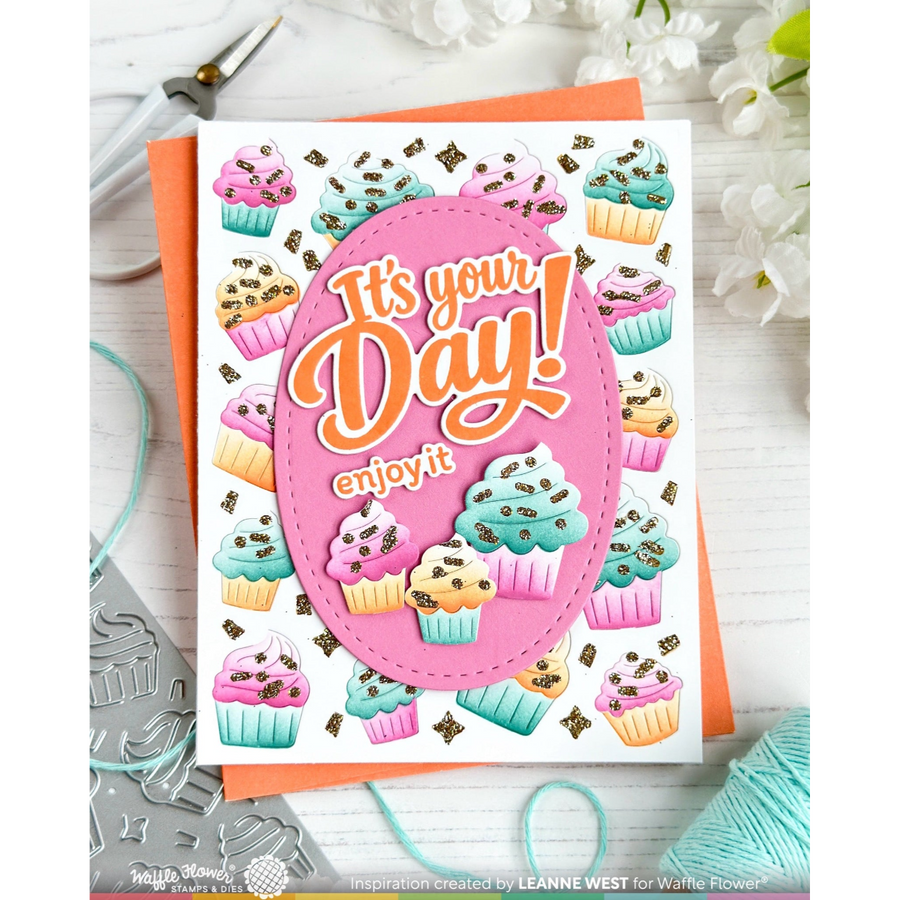 A handmade greeting card featuring colorful cupcakes and the text 'It's your Day! enjoy it' inside a pink oval, displayed on an orange cardstock sheet with crafting tools and yarn around it.