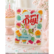 A handmade greeting card featuring a heart-shaped design with the words 'It's your Day!' in red script and 'just for you' on a blue banner, surrounded by colorful 3D cupcake decorations. The card is styled on a white wooden surface with pink cupcakes and flowers in the background.
