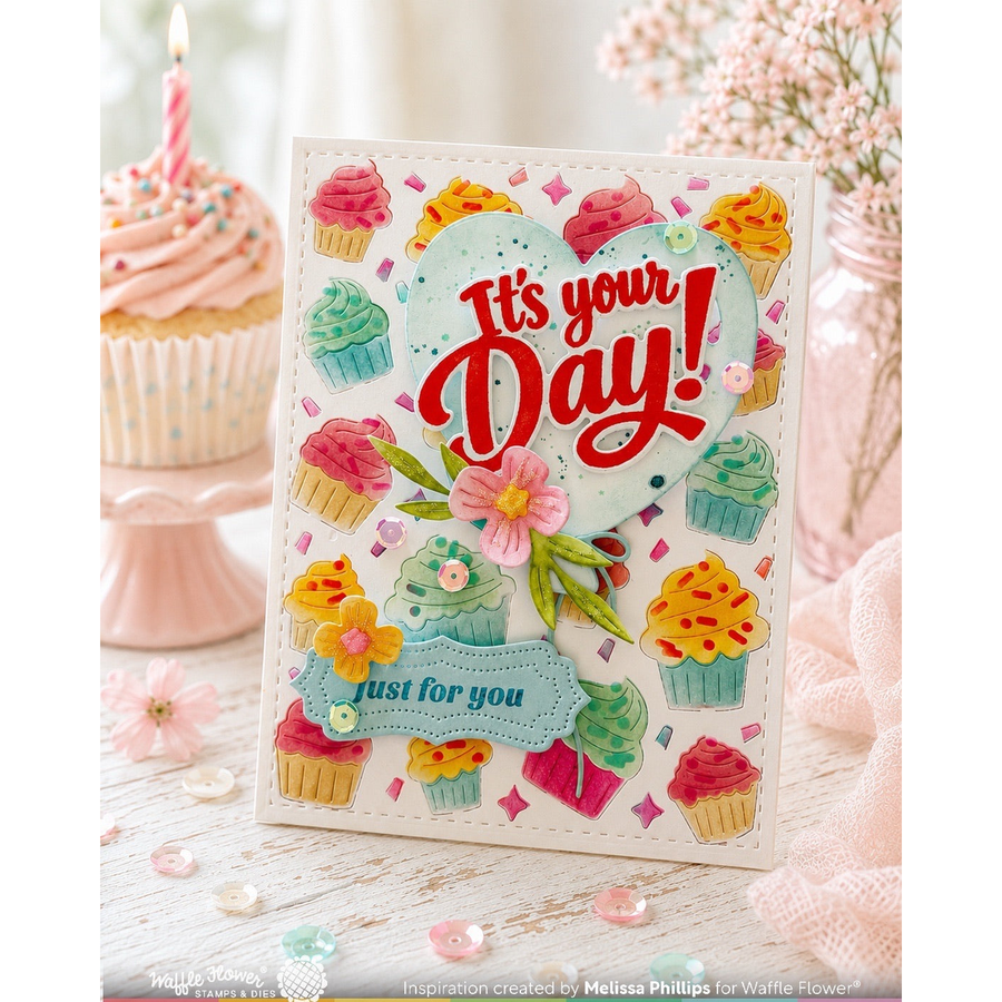 A handmade greeting card featuring a heart-shaped design with the words 'It's your Day!' in red script and 'just for you' on a blue banner, surrounded by colorful 3D cupcake decorations. The card is styled on a white wooden surface with pink cupcakes and flowers in the background.