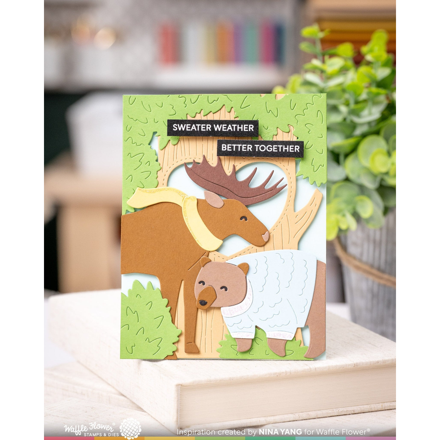 A handmade layered paper card featuring a moose, bear, and a white sweater, with the phrases 'SWEATER WEATHER' and 'BETTER TOGETHER' on a light wood surface with a blurred indoor background.
