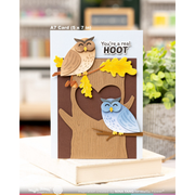 A handmade A7 card featuring two paper owls perched on a tree branch with a heart-shaped tree trunk. The card is displayed on a stack of books, with a blurred background showing a cozy indoor setting.