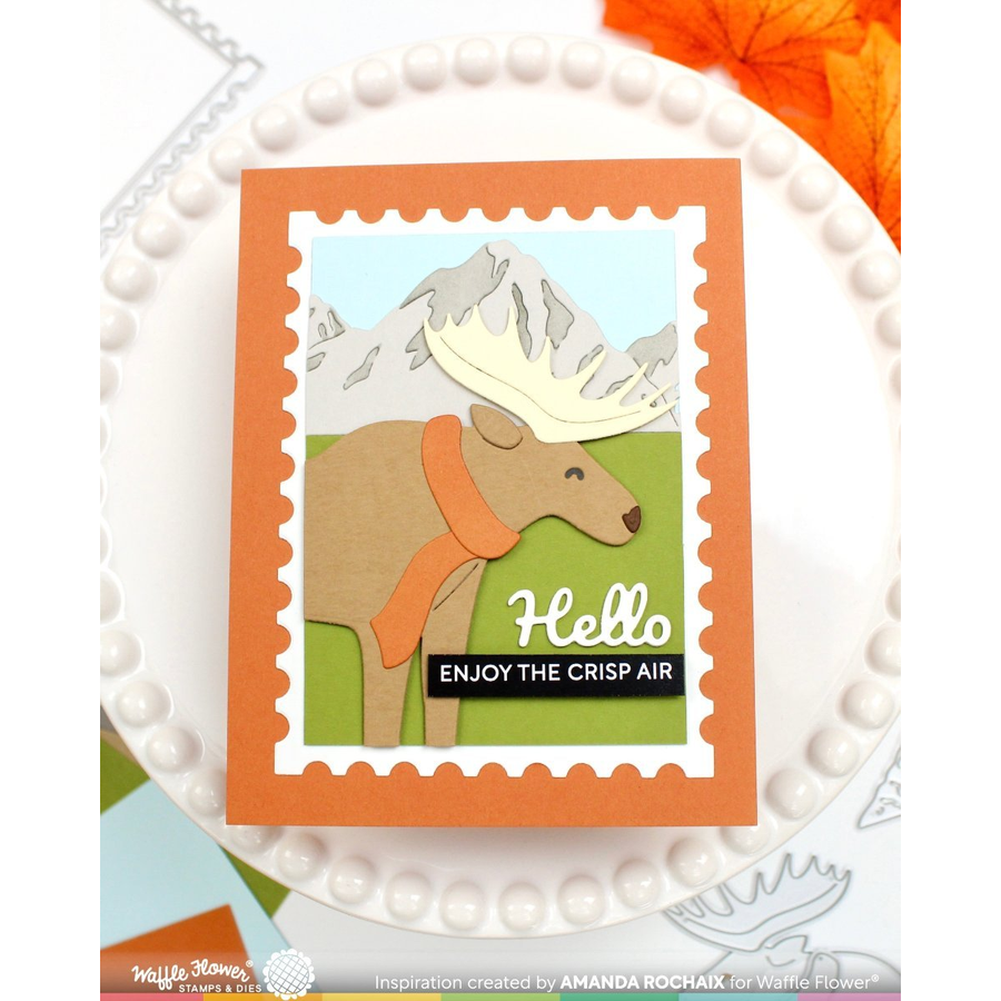 A handmade layered card featuring a die-cut moose wearing an orange scarf against a mountain background, styled on a white beaded plate with an orange leaf in the corner.