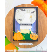 A handmade Halloween greeting card featuring a cute ghosted dog, pumpkins, bats, and a moon, displayed on a wooden coaster with autumn leaves in the corner.