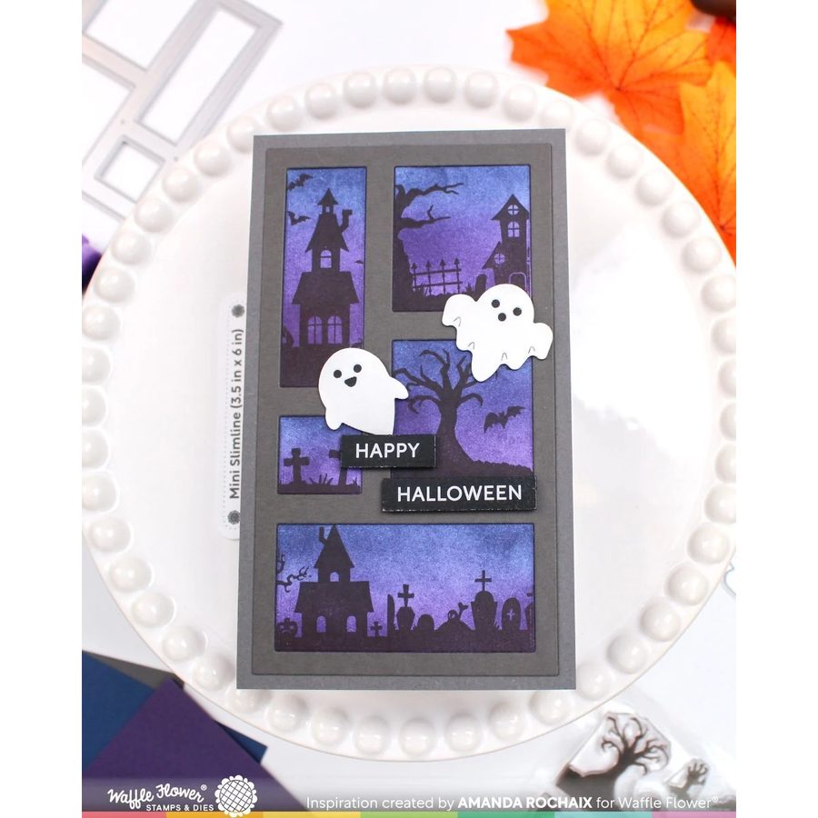A handmade Halloween greeting card with a gray border and four purple-toned haunted house panels, decorated with white ghost cutouts, a black tree silhouette, and the words 'HAPPY HALLOWEEN' on a black banner. The card is displayed on a white beaded plate with autumn leaves and crafting props around it.