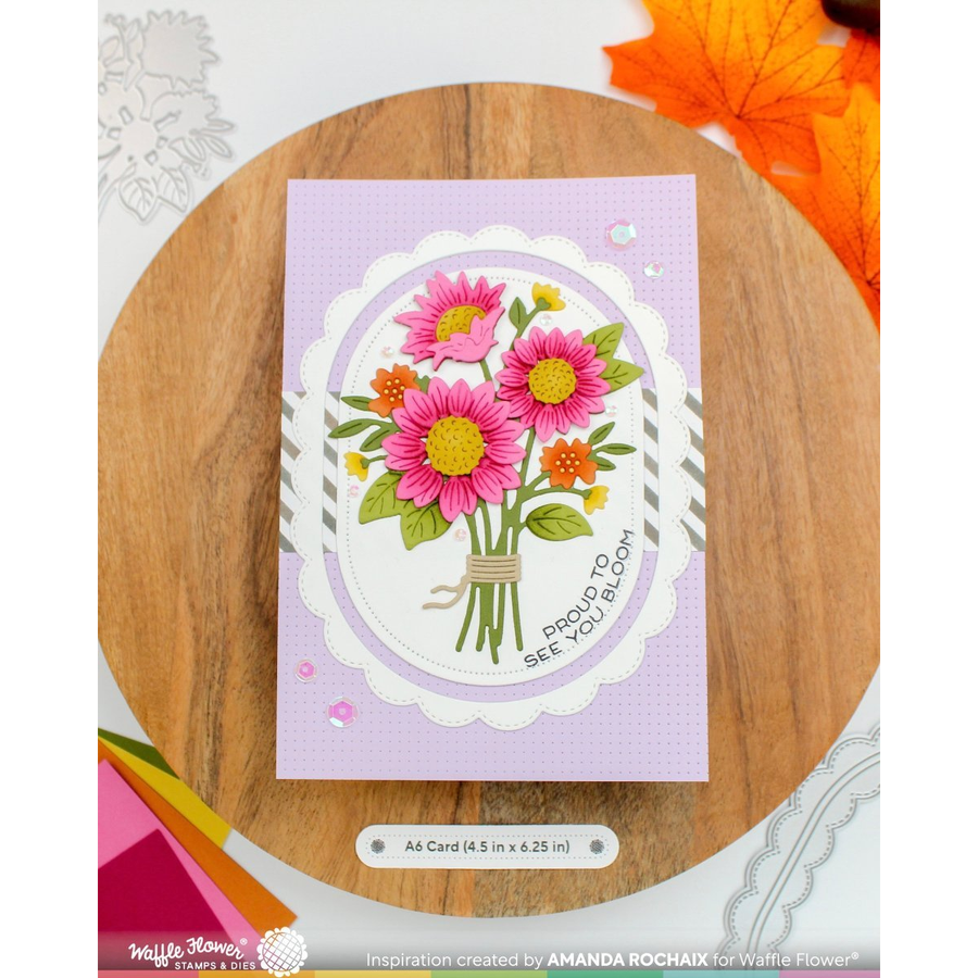 A handmade greeting card featuring a bouquet of pink and orange daisies with green leaves, displayed on a round wooden board. The card has a light purple border and the text 'PROUD TO SEE YOU BLOOM'.