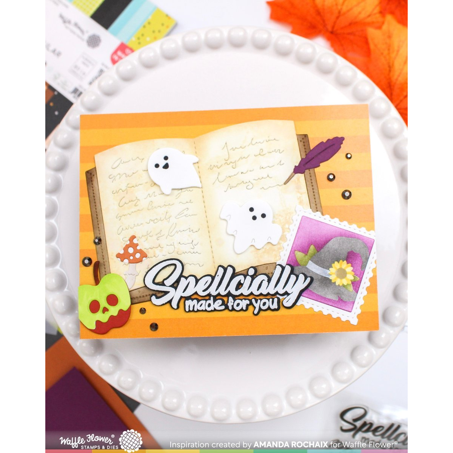 A handmade greeting card featuring a ghost-themed Halloween design, displayed on a white beaded plate. The card has a warm orange background, an open book with handwritten script, white ghost cutouts, a purple feather, a small pumpkin, a green skull, and a purple postage stamp graphic.