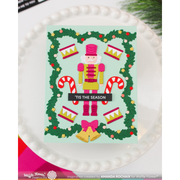 A handmade Christmas card featuring a nutcracker surrounded by a green wreath with candy canes, bells, and bows, displayed on a white plate. The card has a mint green background and a black banner reading 'TIS THE SEASON'.