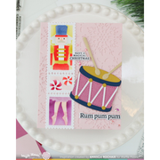 A handmade Christmas card featuring a pink background with a nutcracker, a pink-and-yellow drum, candy canes, and the text 'HAVE A MAGICAL CHRISTMAS' and 'Rum pum pum', displayed on a white plate.