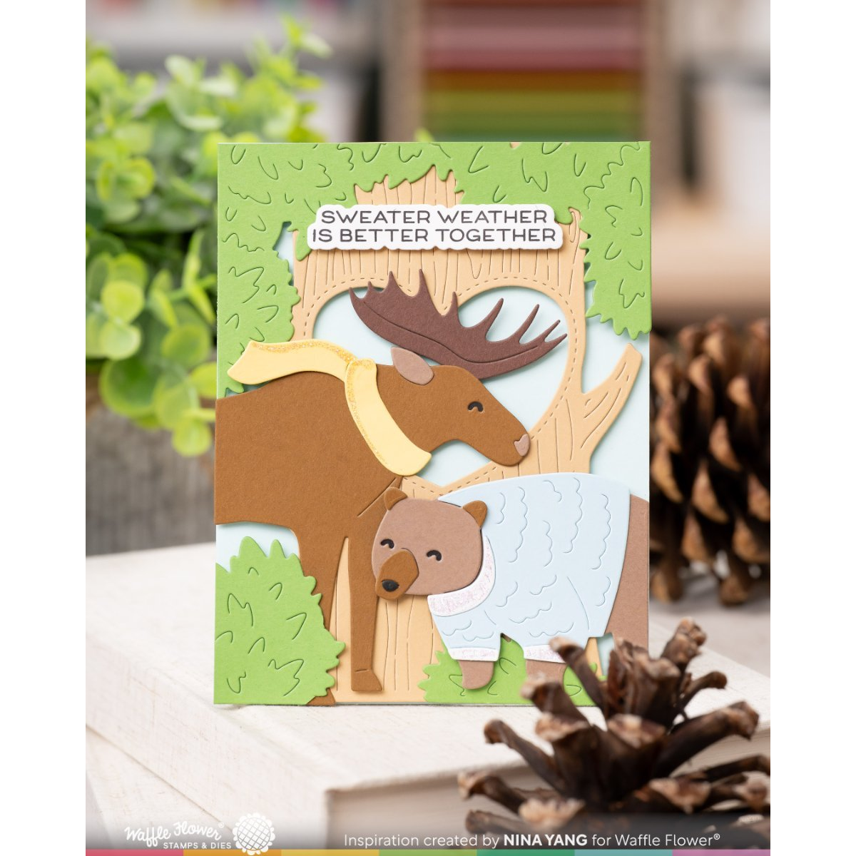 A handmade layered card featuring a brown moose and a bear in a forest scene, with the text 'SWEATER WEATHER IS BETTER TOGETHER' on a white banner. The card is displayed on a wooden surface with blurred pinecones and greenery in the background.