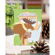 A handmade layered card featuring a brown moose and a bear in a forest scene, with the text 'SWEATER WEATHER IS BETTER TOGETHER' on a white banner. The card is displayed on a wooden surface with blurred pinecones and greenery in the background.