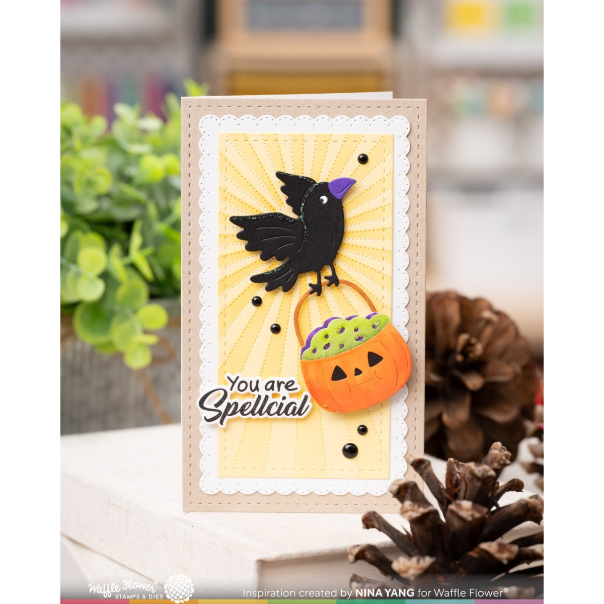 A handmade greeting card featuring a black raven holding a jack-o'-lantern bucket with purple and green candy, set against a yellow sunburst background with the text 'You are Spellcial'. The card is styled on a shelf with pinecones and greenery in a blurred background.