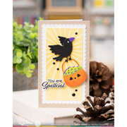 A handmade greeting card featuring a black raven holding a jack-o'-lantern bucket with purple and green candy, set against a yellow sunburst background with the text 'You are Spellcial'. The card is styled on a shelf with pinecones and greenery in a blurred background.