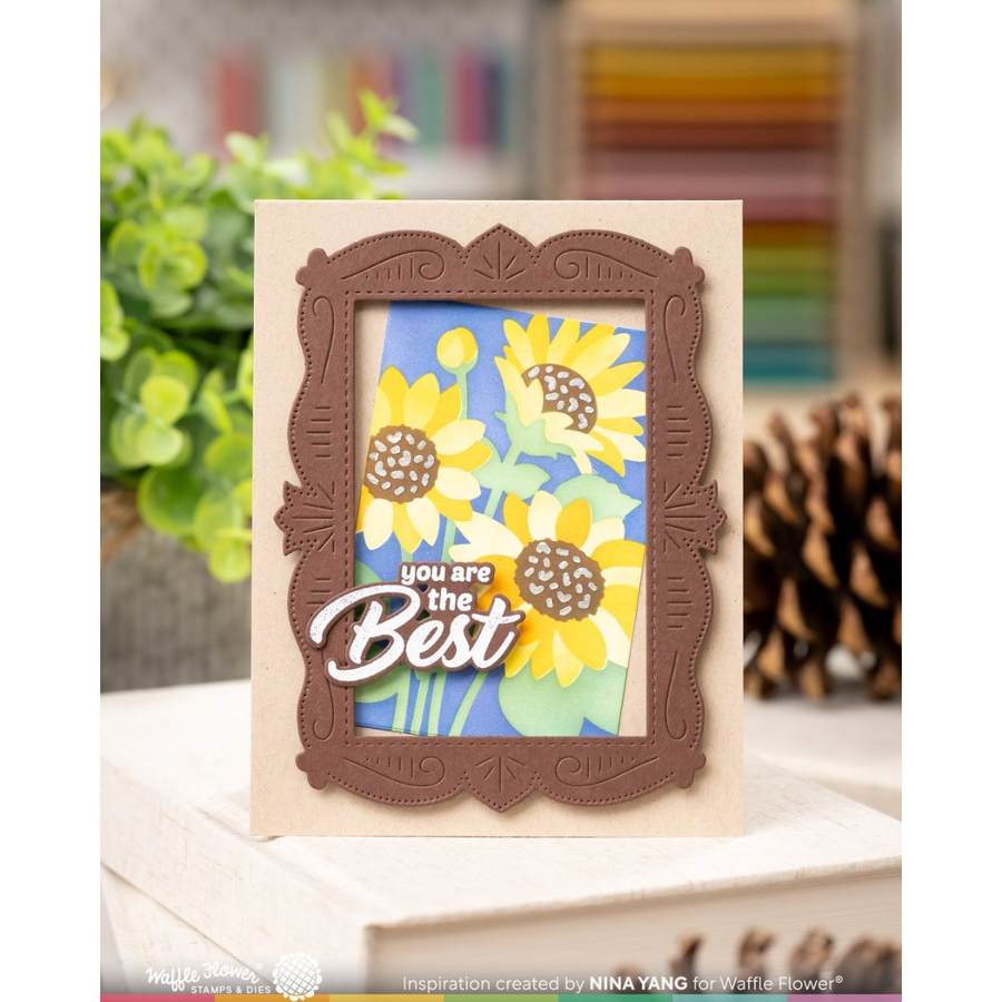 A handmade greeting card featuring a brown ornate frame around a layered sunflower design on a blue background, with the phrase 'you are the Best' in white script. The card is displayed on a wooden surface with a blurred background of greenery and books.