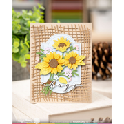 A handmade greeting card featuring three yellow sunflowers with brown centers, green leaves, and a white scalloped oval background, layered over a tan burlap-textured design. The card is displayed on a wooden surface with a blurred background of greenery and a pine cone.