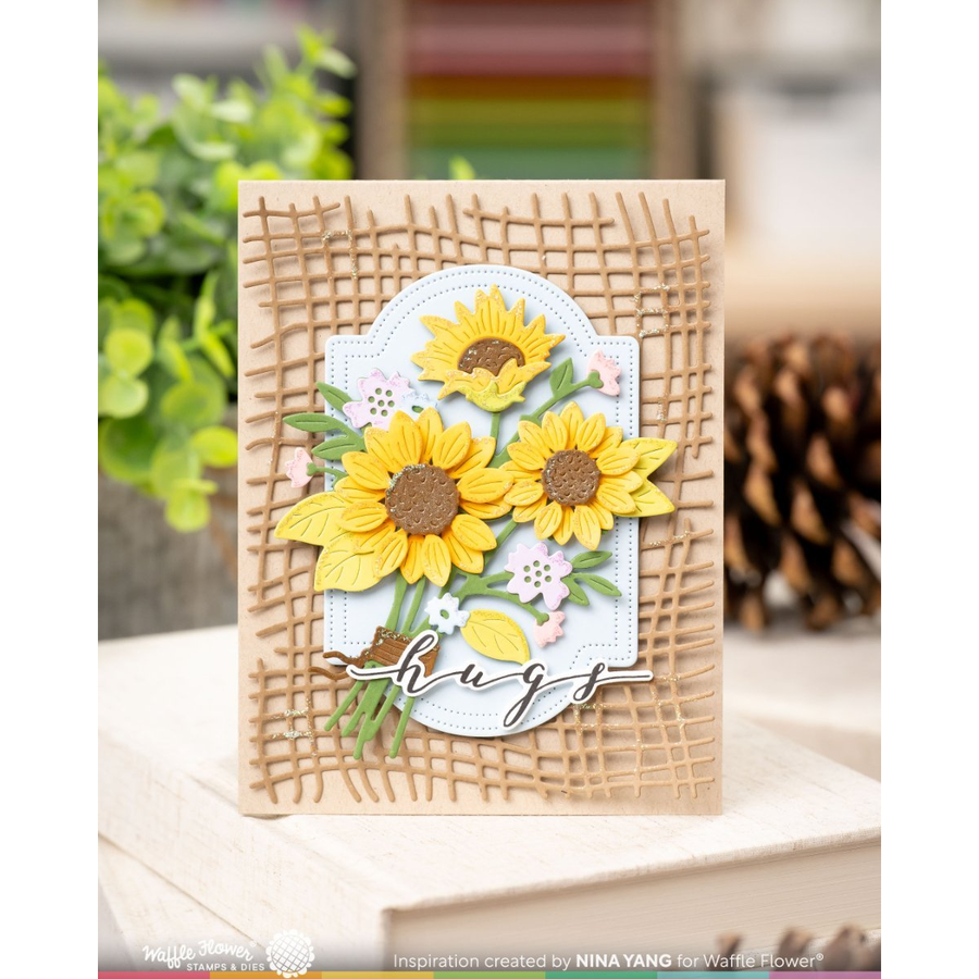 A handmade greeting card featuring three yellow sunflowers with brown centers, green leaves, and a white scalloped oval background, layered over a tan burlap-textured design. The card is displayed on a wooden surface with a blurred background of greenery and a pine cone.