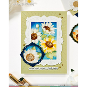 A handmade A6 greeting card featuring layered die-cut floral designs, including white daisies with brown centers and yellow sunflowers on a blue background, set on a light olive green card base. The card includes a scalloped white border, a dark blue oval accent, and the printed phrase 'SENDING LOVE YOUR WAY' at the bottom.