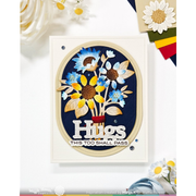 A handmade greeting card featuring a circular floral design with sunflowers and daisies on a navy blue background, with the text 'Hugs' and 'THIS TOO SHALL PASS'. The card is displayed on a white surface with decorative daisy flowers and craft paper accents.