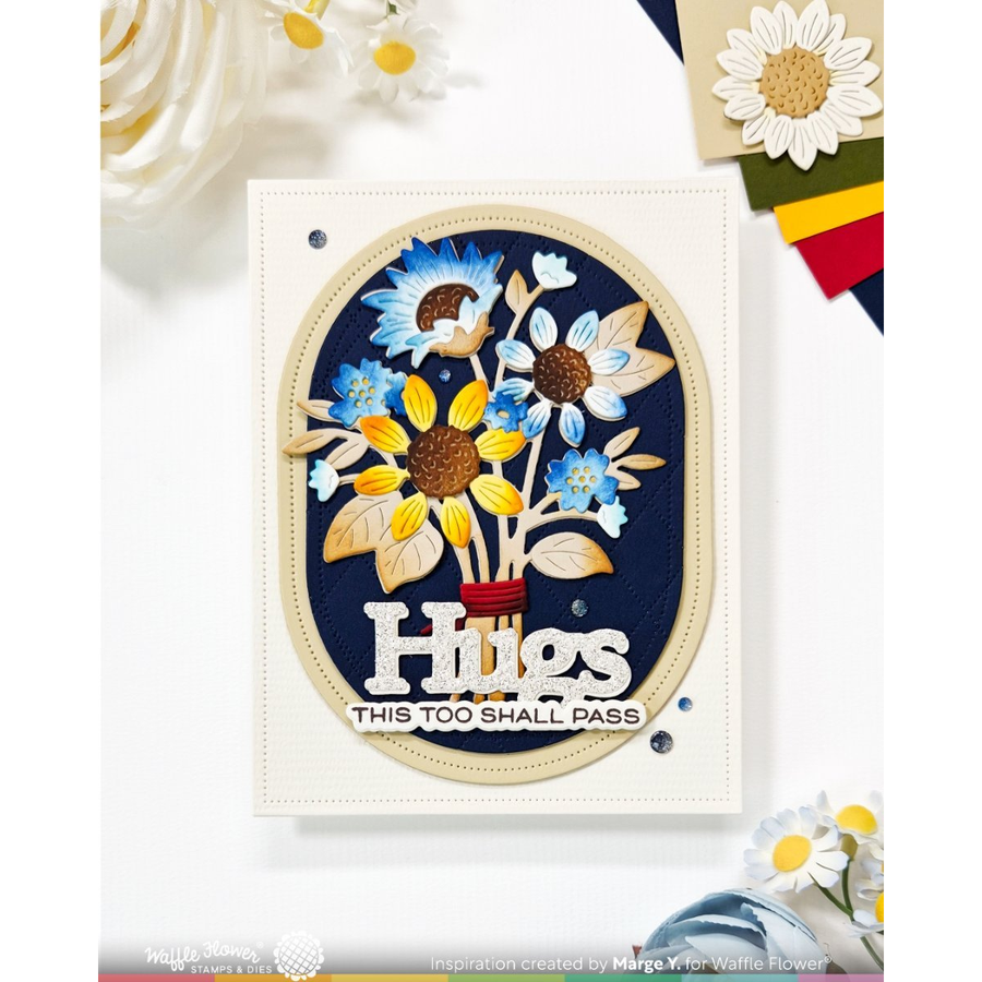 A handmade greeting card featuring a circular floral design with sunflowers and daisies on a navy blue background, with the text 'Hugs' and 'THIS TOO SHALL PASS'. The card is displayed on a white surface with decorative daisy flowers and craft paper accents.