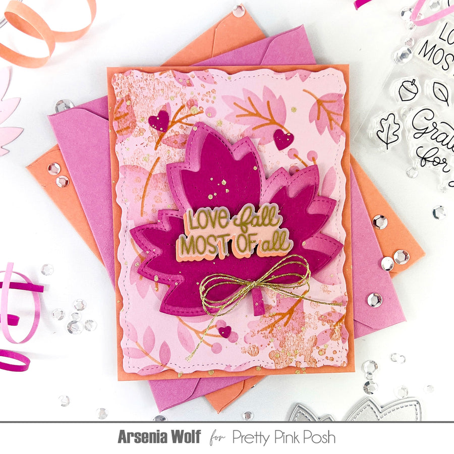 A handmade layered card featuring autumn leaf designs in shades of pink, magenta, and orange, with a gold-foiled 'LOVE fall MOST OF all' sentiment and a small gold bow. The card is displayed on top of matching pink and orange envelopes on a white background decorated with craft elements.