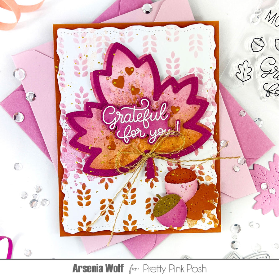 A handmade layered card featuring a large pink and orange autumn leaf with the phrase 'Grateful for you!' in white script, surrounded by pink envelopes, acorns, and decorative embellishments on a white background. Text at the bottom reads 'Arsenia Wolf for Pretty Pink Posh'.