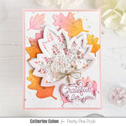 A handmade layered card featuring a white maple leaf with pink and orange glitter accents, sequins, and a small gold bow, surrounded by colorful die-cut autumn leaves. The card includes a pink script tag that reads 'Grateful for you!' and is photographed on a white wood surface with decorative flowers in the background.