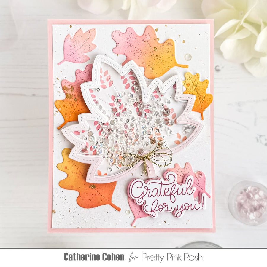 A handmade layered card featuring a white maple leaf with pink and orange glitter accents, sequins, and a small gold bow, surrounded by colorful die-cut autumn leaves. The card includes a pink script tag that reads 'Grateful for you!' and is photographed on a white wood surface with decorative flowers in the background.
