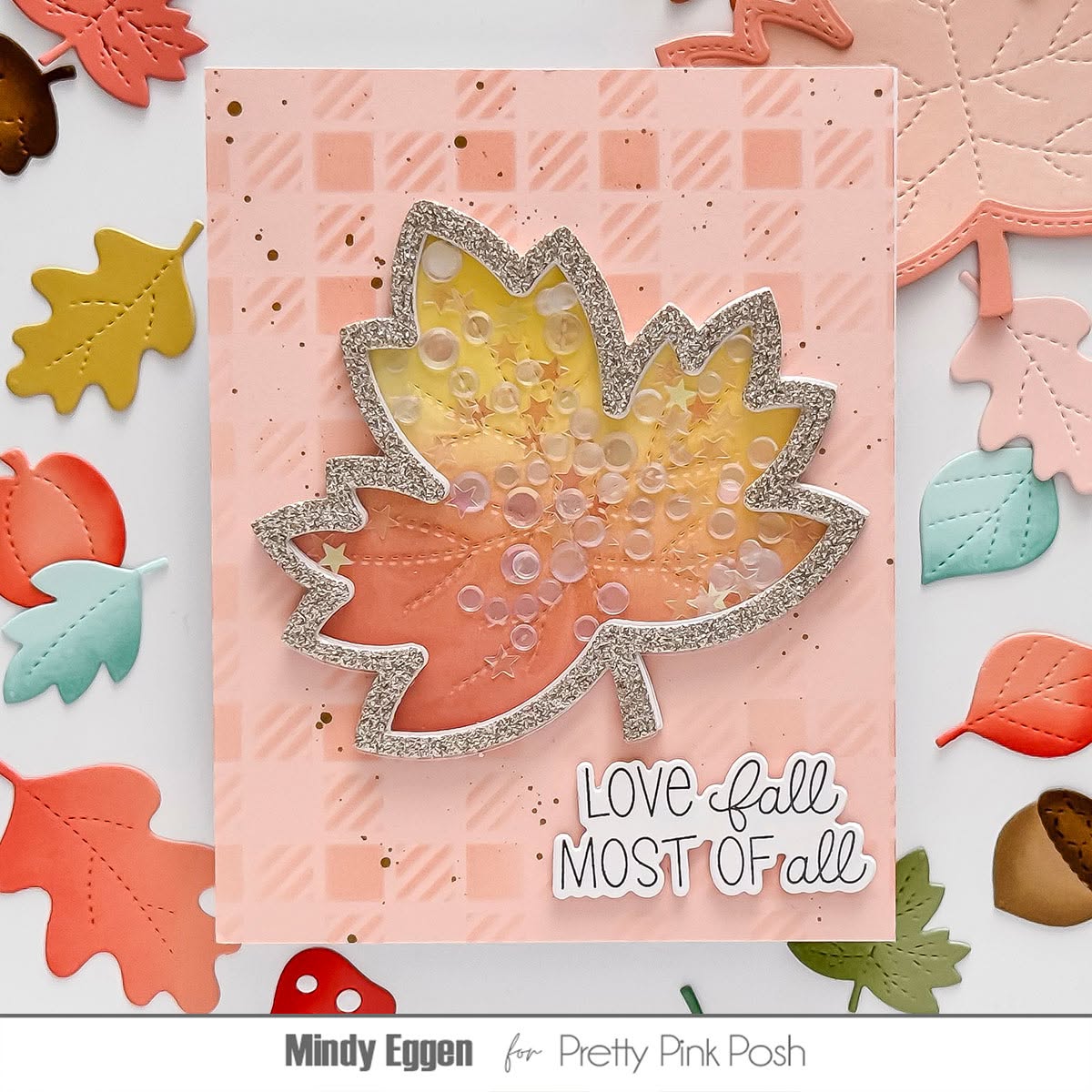 A handmade autumn-themed card featuring a glittery silver maple leaf with a yellow, orange, and pink gradient center filled with clear bubble elements. The card has a pink checkerboard patterned background and the text 'LOVE fall MOST OF all'.