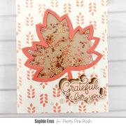 A handmade greeting card featuring a large maple leaf with a transparent center filled with multicolored sequins, surrounded by a coral-pink stitched border. Below the leaf, the words 'Grateful for you!' are written in metallic gold script.